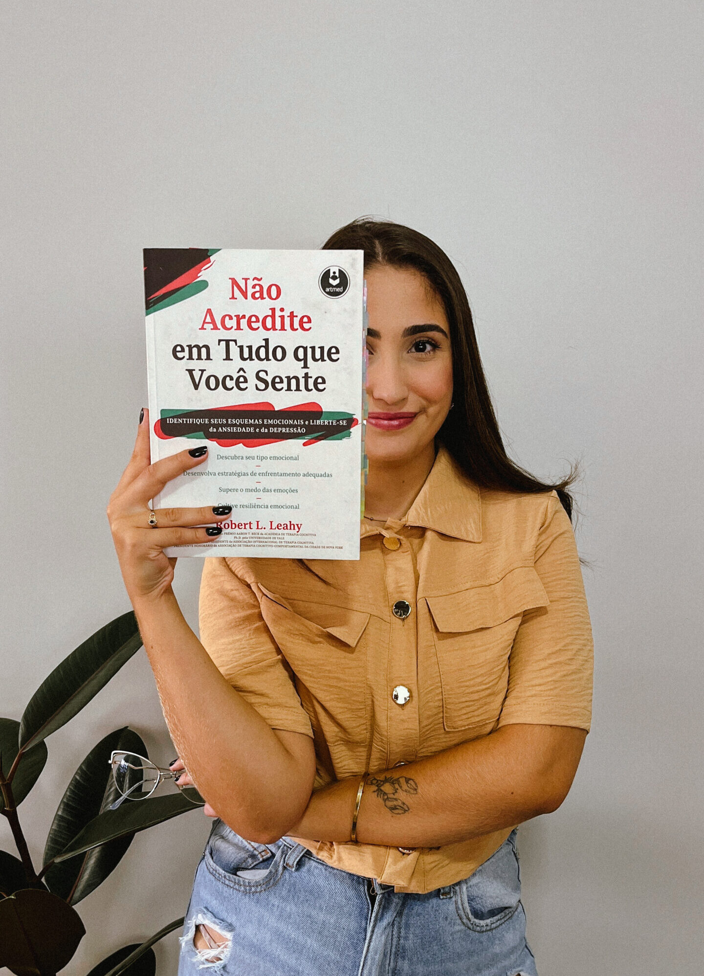 Foto de Luana morais em pé segurando um livro com o título "Não acredite em tudo que você sente" em frente a metade do rosto